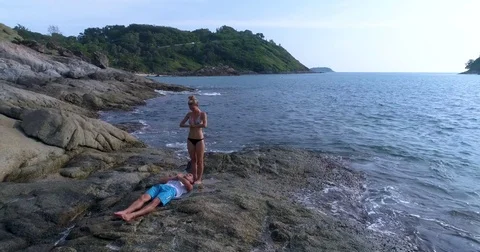 Aerial: A beautiful young couple is practicing yoga on the rocks next to the sea Stock Footage 82189126