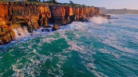 Aerial drone view of turquoise ocean waves crashing against rugged golden cliffs Stock Footage 345293046