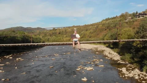 Aerial drone view of young man in white shirt sitting on wooden bridge over Stock Footage 165527434