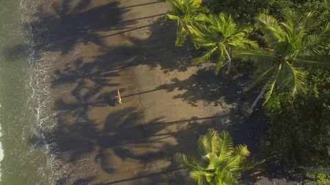 AERIAL: Muscular guy holding surfboard and walking along sandy beach in Bali Stock Footage