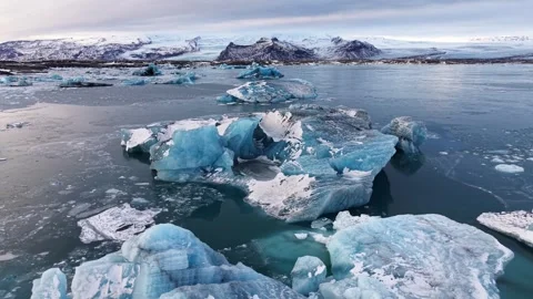 Aerial pullback over icebergs with snowy mountains at sunrise in glacier lag Video stock 327823325