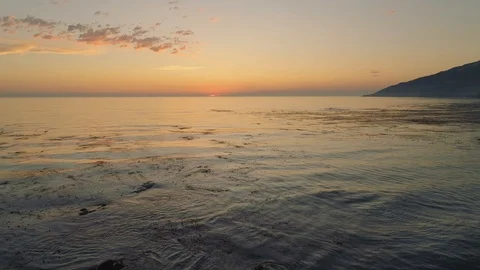 Aerial shot flying backwards looking at a sunset by the ocean. Slowly rising. Video stock 87728392