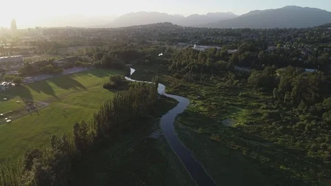 Aerial Shot Flying Over A River Besides Sports Fields With Mountains Behind Stock Footage 100532001