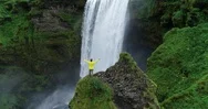 Aerial Shot Of Man With Arms Raised Standing In Front Of Waterfall Celebrating Stock Footage