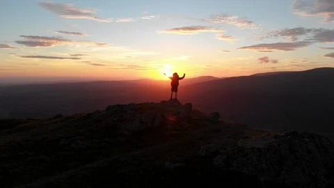 Aerial shot of a man celebrating at the top of a mountain at sunset Stock Footage