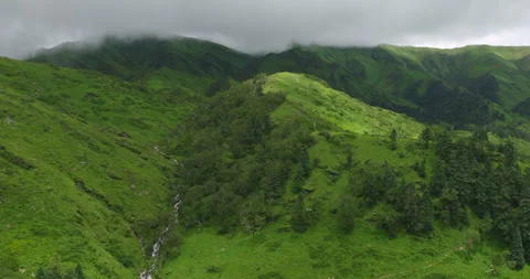 Aerial shot over mountain stream through lush alpine ridge and foggy peaks Stockbeeldmateriaal 346797153