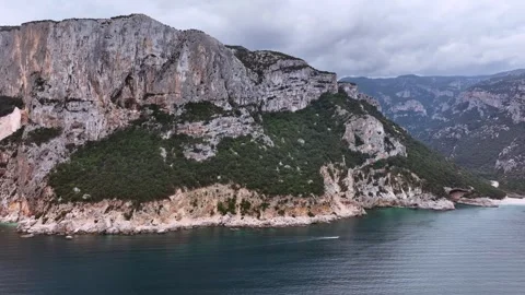 Aerial shot of a tiny boat sailing past a massive cliff. Orosei coast Stock Footage 260618587
