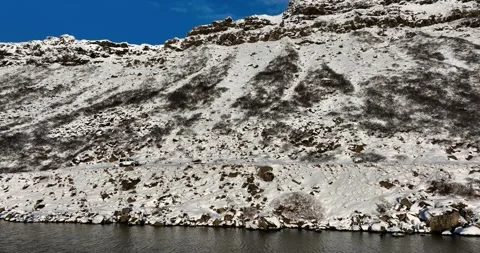 Aerial side view of a white 4x4 moving along the snowy cliffside shore of 動画素材 334376733