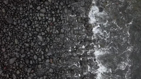Aerial Top Down: Dark Smooth Pebbles and White Sea Foam on Rocky Shore. Video stock 310115534