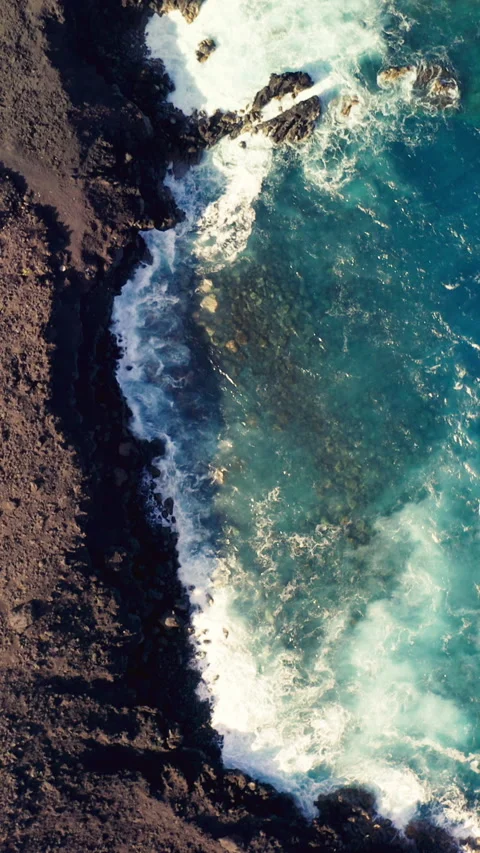 Aerial top-down view of turquoise waves breaking against black volcanic cliffs Stock Footage 320947649