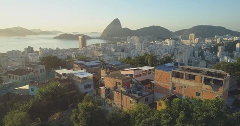 Aerial tracking behind mountaintop favela in forest at sunrise with high rise 스톡 동영상 101730562