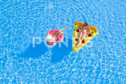 AERIAL: Two cheerful girlfriends lying on inflatable floaties, relaxing in pool Stock Photos
