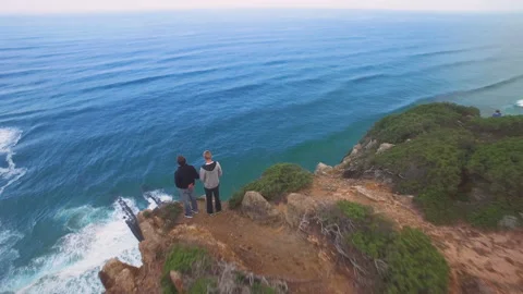 Aerial of two men standing on cliff with ocean waves splashing against rocks Stock Footage 158636979