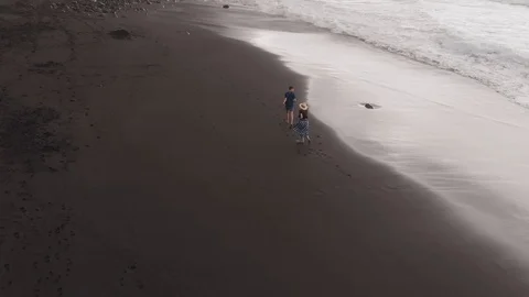 Aerial view of black beach. A man and a woman run fast on the volcanic sand Видео 112216165