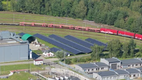 Aerial view of a cargo train passing a solar panel farm and industrial complex 스톡 동영상 348270900