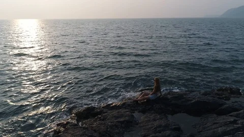 Aerial view. Flying around a young girl sitting on edge of a stone reef by sea. Vídeos de archivo 104465945