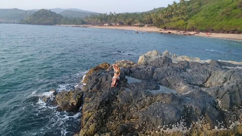 Aerial view. Flying around a young girl sitting on edge of a stone reef by sea. 스톡 동영상 104469167