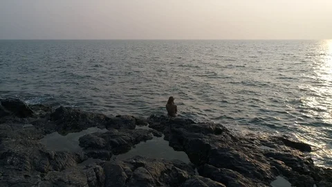 Aerial view. Flying around a young girl sitting on edge of a rocky reef by sea. Stock Footage 104473953