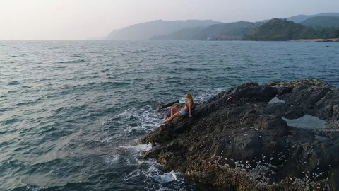 Aerial view. Flying around a young girl sitting on edge of a stone reef by sea. Stock Footage 104476007