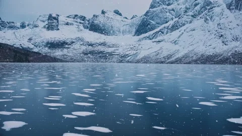 Aerial view from low flying drone of frozen lake in snowfall. Mountains in wi Stock Footage 295544395