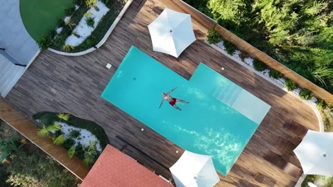 Aerial view of man floating in luxurious backyard swimming pool with wooden deck Stock Footage 301785800