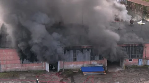 Aerial view of massive industrial fire with thick black smoke and fire trucks Stock Footage 321362697