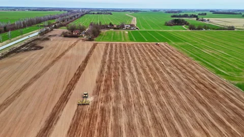 Aerial view of multiple tractors preparing a large brown field next to a lush Stock Footage 319623957