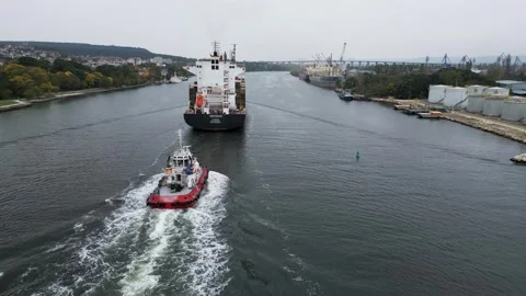 Aerial view of a red tugboat escorting a large cargo ship through the industrial Stock Footage 319749976