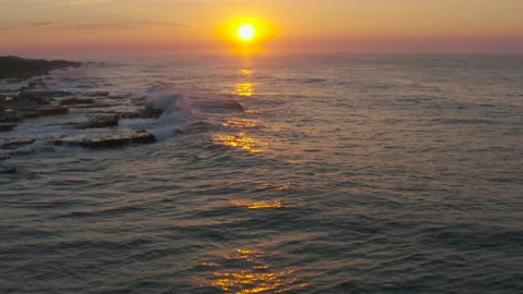 Aerial view of the setting sun and waves breaking on the rocks of a coastline Stock Footage 153759634