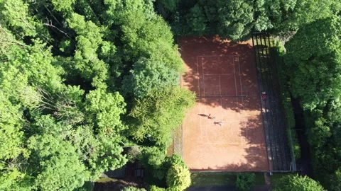 This aerial view shows a red clay tennis court surrounded by dense green trees. 스톡 동영상 336632802