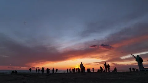 Aerial View: Silhouettes of people enjoy a calm moment, gazing at the orange sun Stock-Footage 296162653