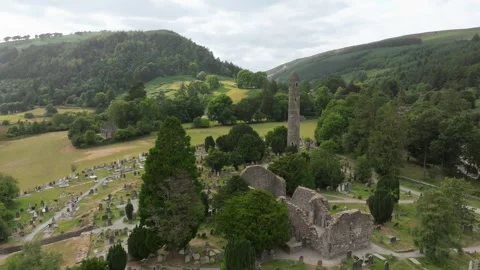 Aerial View of Stone Round Tower, Cemetery and Church Ruins in a Wooded Valley Stock Footage 348742638