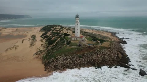 Aerial view of a tall white lighthouse perched on a rocky hill by the sea. Stock Footage 297635853