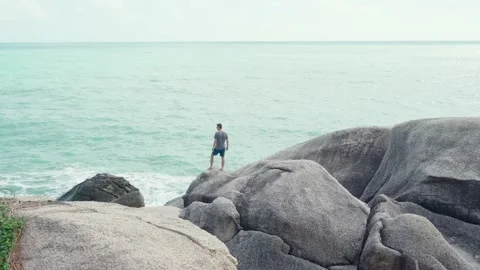 Aerial view of Young strong man standing on big rocks on the coast of ocean. Stock Footage 219508574