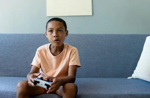 African American boy playing while holding white and black controller on sofa in Stock Photos
