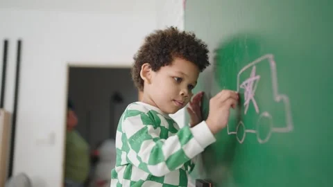 African american boy using a chalk drawing on chalk board in classroom at school Stock Footage 248545527