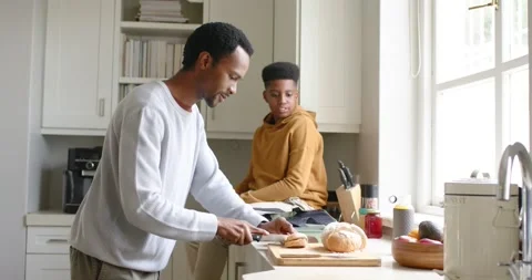 African American dad slicing bread for breakfast in kitchen as teenage son Stock Footage 337745543