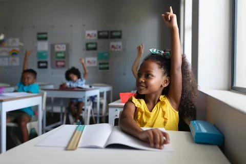 African american elementary school students with hands raised sitting at desk in Stock Photos