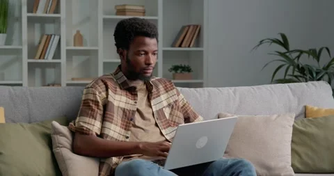African American guy in checked shirt sitting on sofa while using laptop. Stock Footage 249465871