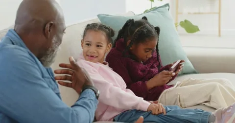 African American man and two girls gesturing on sofa older tapping phone drawing Stock Footage 346178724