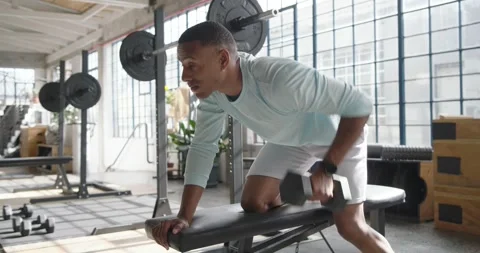 African American man in blue top picking up dumbbell, performing rows on loft Stock Footage 338338341