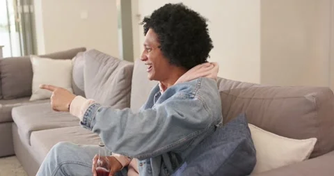 African American man with bottle on sofa reacting to offscreen sight laughing Stock Footage 341320159