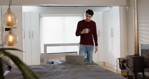 African American man checking laptop, standing behind bed holding ceramic mug Stock Footage 339722140