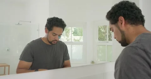 African American man checking mirror, applying two blue eye gel patches at sink 動画素材 334843313