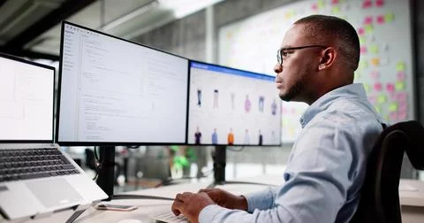 African American Man Coding Ecommerce Software On Multiple Office Monitors. Stock Photos