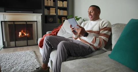 African American man on couch by fireplace in white sweater reading tablet Stock Footage 342363552