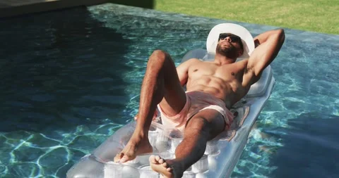 African American man lying on clear lounger in pool, white hat, sun glinting, Stock Footage 344334052