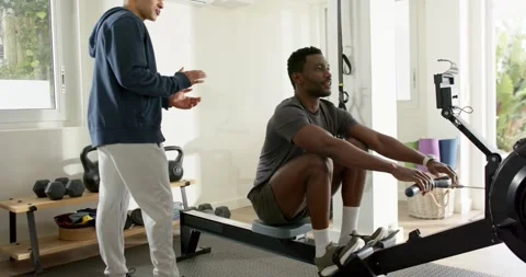 African American man rowing on machine after trainer pointing monitor for Stock Footage 337705053