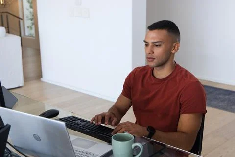African American man in rust-tee typing at home glass desk on keyboard beside Stock Photos