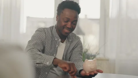 African american man sits on a couch and at a table, putting money in a piggy Stock Footage 282959569